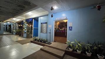 A hotel reception area with a blue wall, a reception desk, and a sign indicating the reception. There are potted plants and a staircase leading to the entrance.