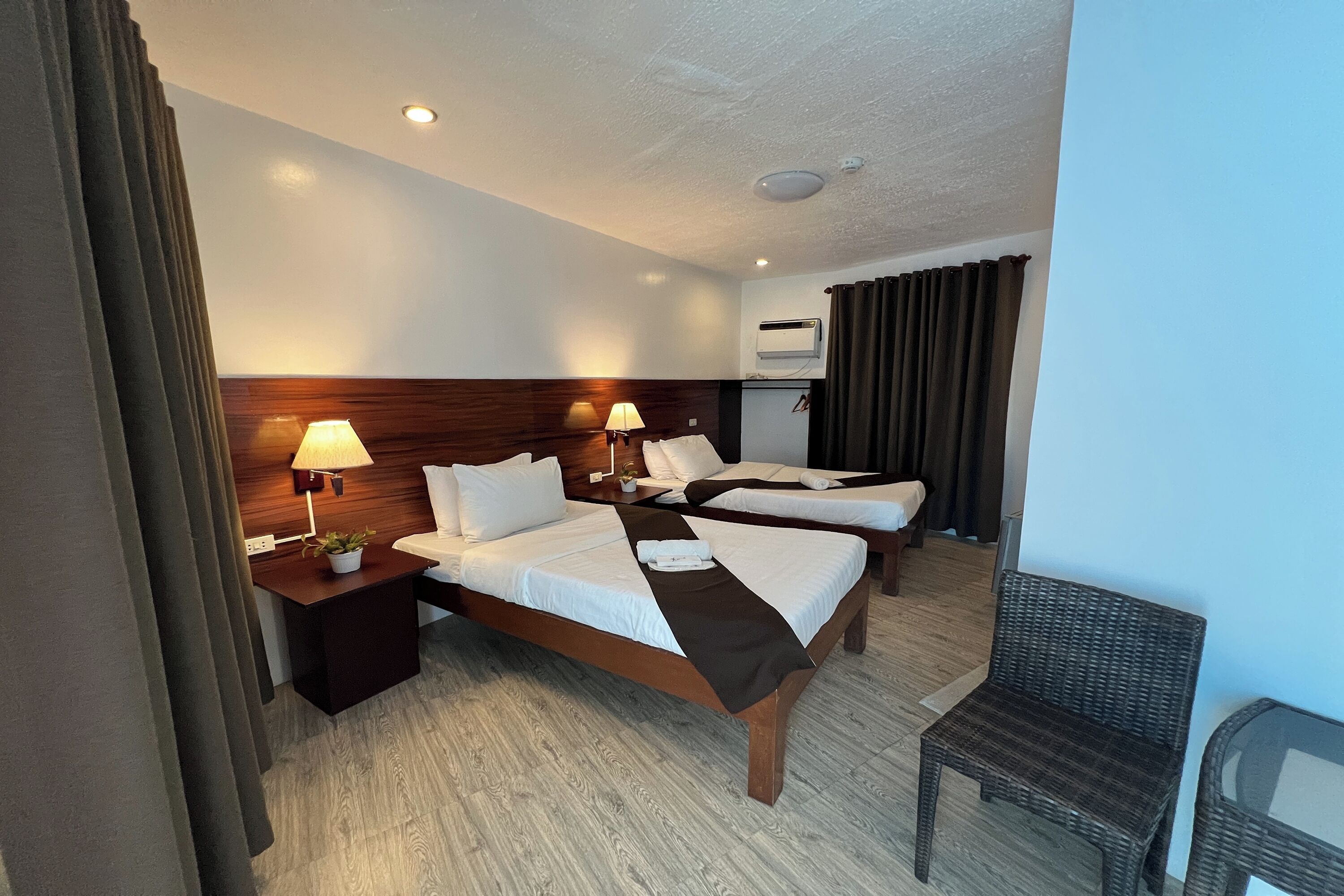 A hotel room with three beds, a wooden headboard, a small table with a lamp, and a wicker chair.