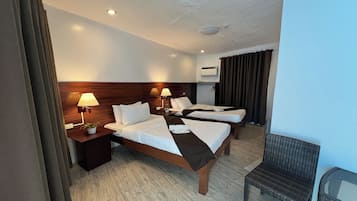 A hotel room with three beds, a wooden headboard, a small table with a lamp, and a wicker chair.