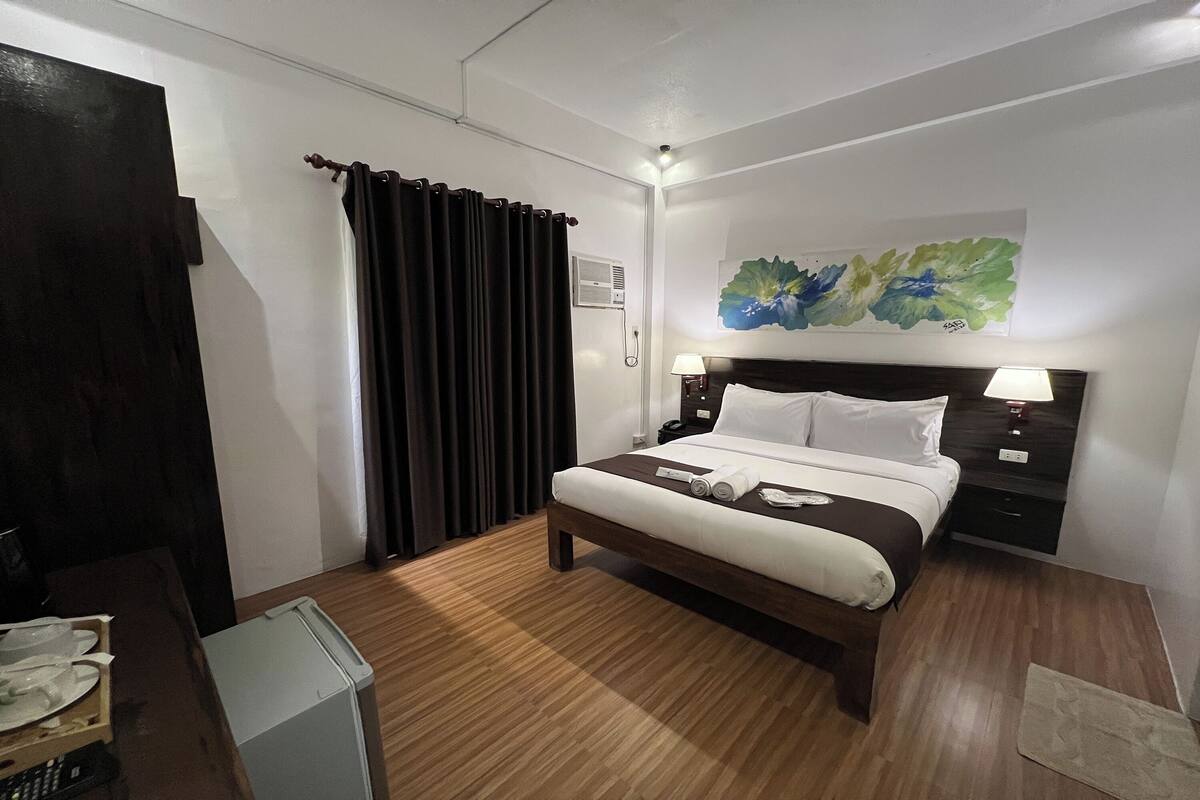 A hotel room with a bed, a nightstand, a wall-mounted air conditioner, and a painting.
