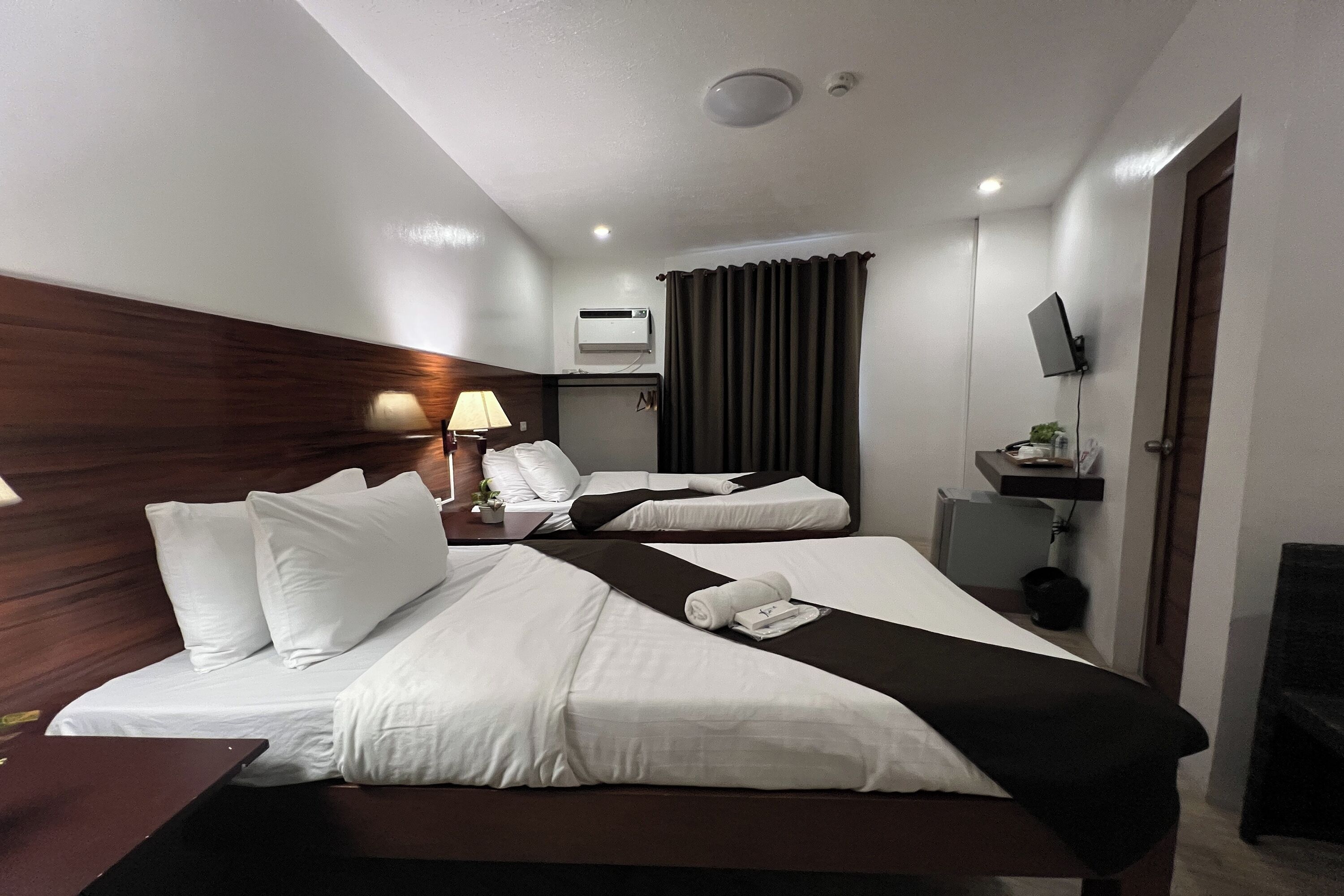 A hotel room with two beds, a desk, a lamp, an air conditioning unit, a TV, and a window with curtains.