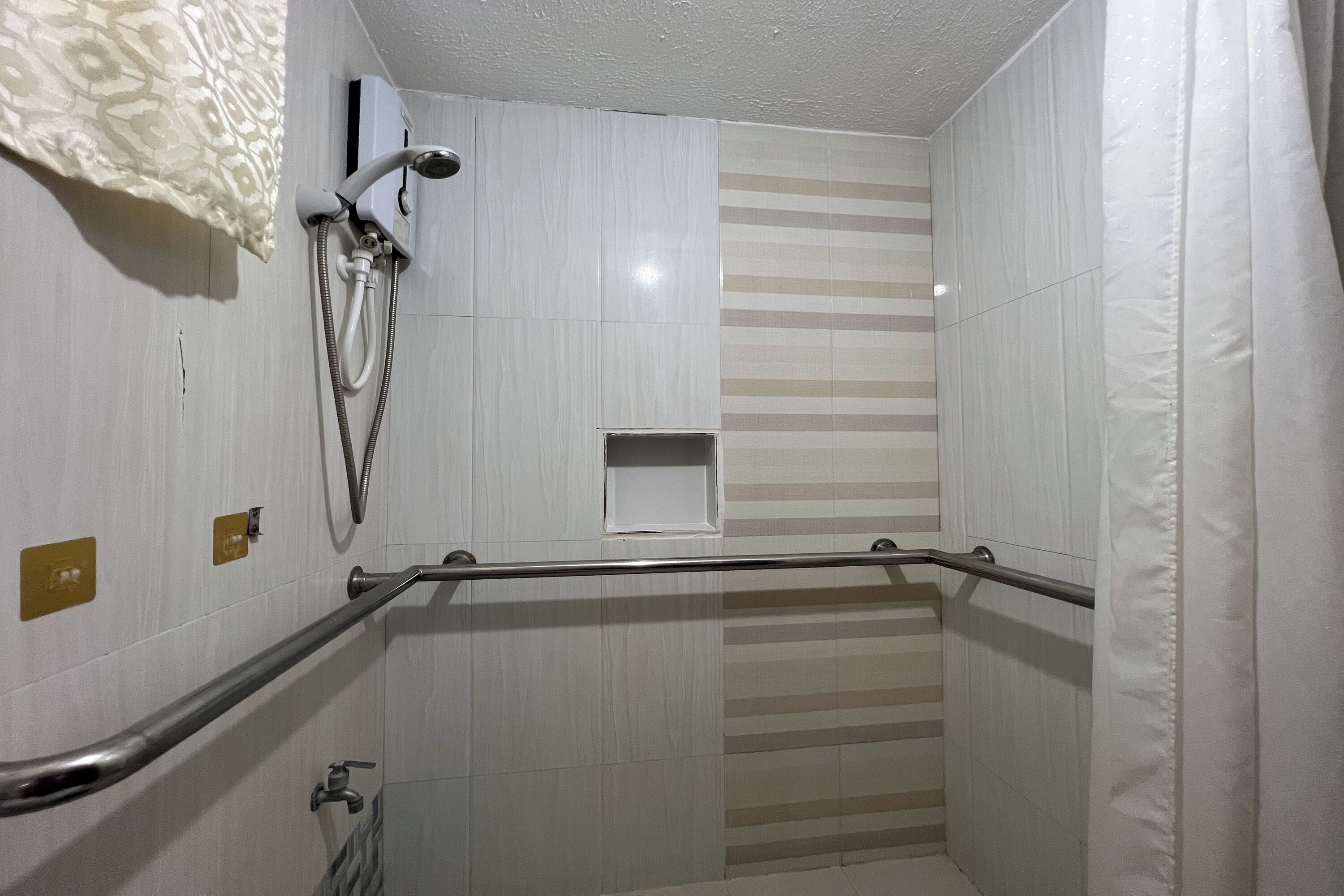 Une salle de bain carrelée de carreaux blancs, une douche avec un pommeau de douche amovible, une barre d’appui et un porte-serviettes.