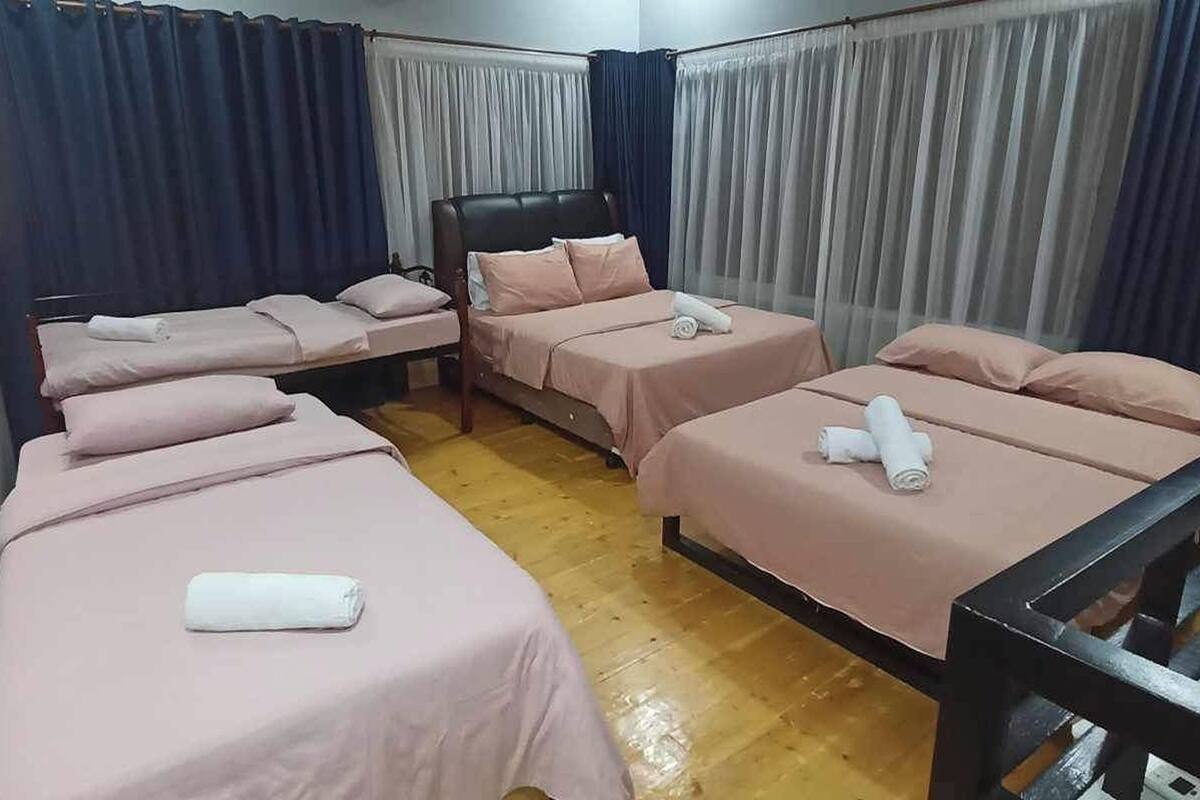 A hotel room with four single beds, each with a pink bedspread and white towels. The room has wooden flooring and is furnished with a black headboard and nightstands.