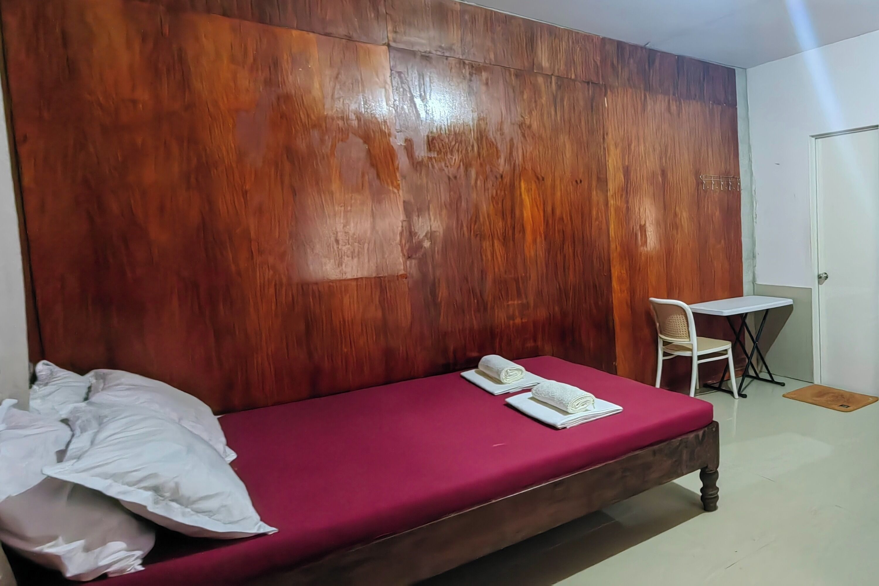 A single bed with a red mattress, a wooden headboard, a white pillow, and two rolled white towels.