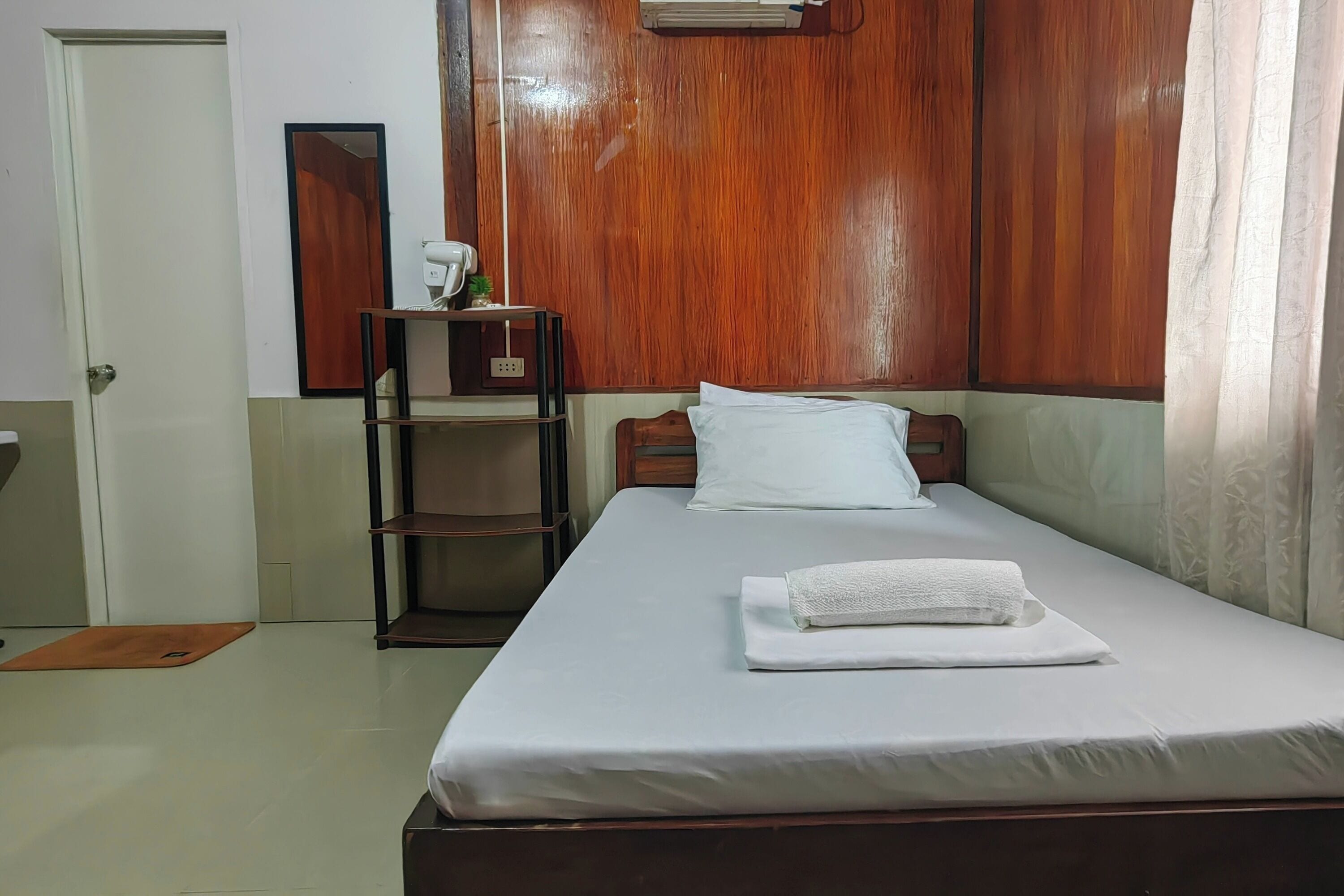 A neatly made bed with a white pillow and a folded towel, a wooden wardrobe, a small shelf with a plant, and a door to the left.