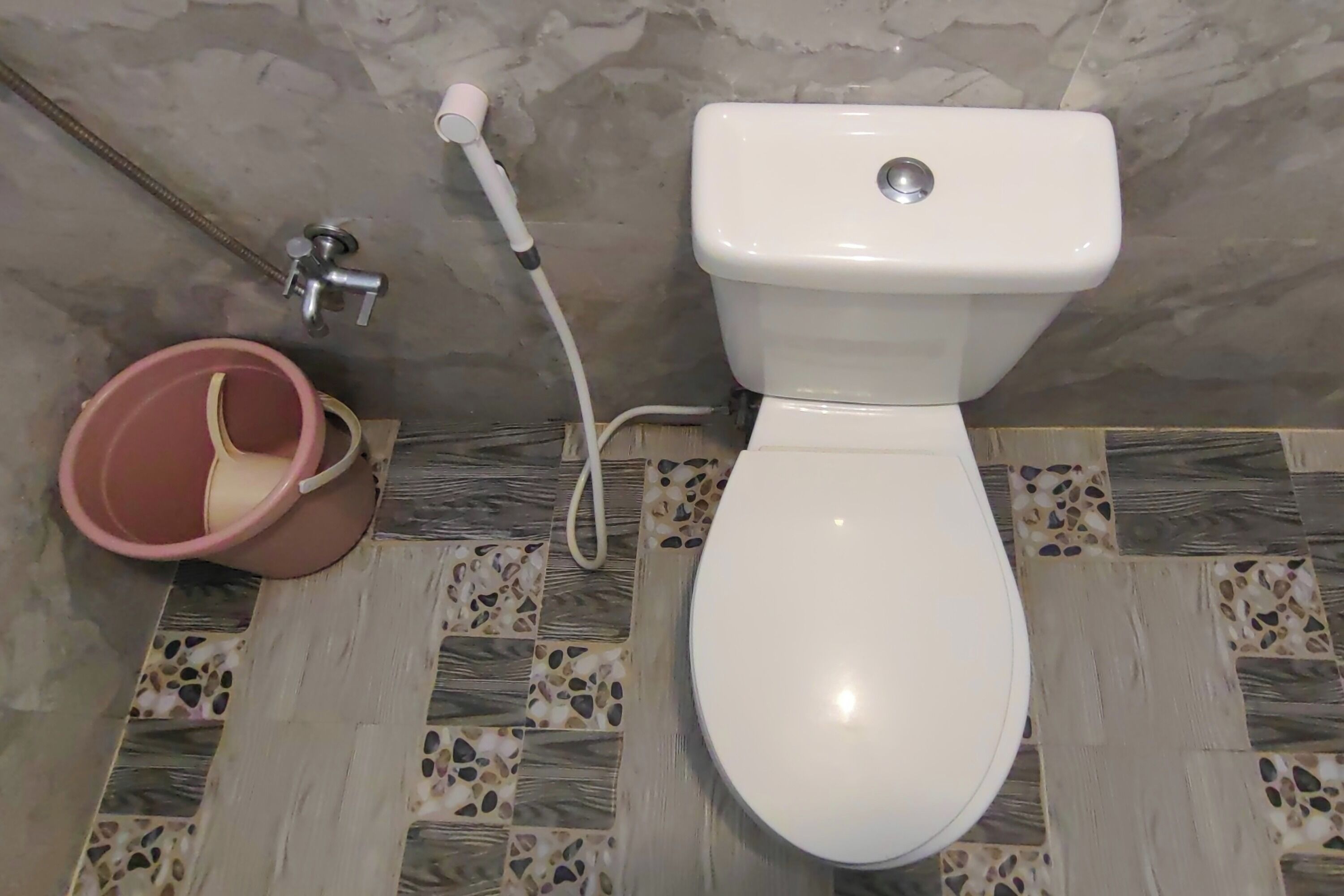 A toilet with a white seat and tank, a pink bucket, and a shower hose.