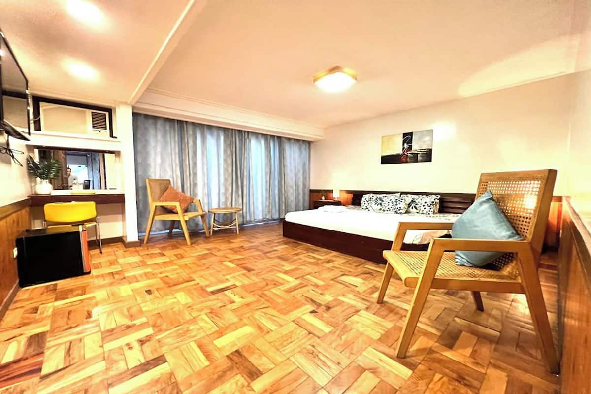 A hotel room with a wooden parquet floor, a bed with a patterned headboard, a sofa with cushions, a small table, a chair, and a desk with a lamp.
