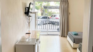 A compact hotel room with a bed, a small desk with a computer and a chair, a window with a view of parked cars and trees, and an air conditioning unit on the wall. - Jommalai Boutique Hotel (Phitsanulok)