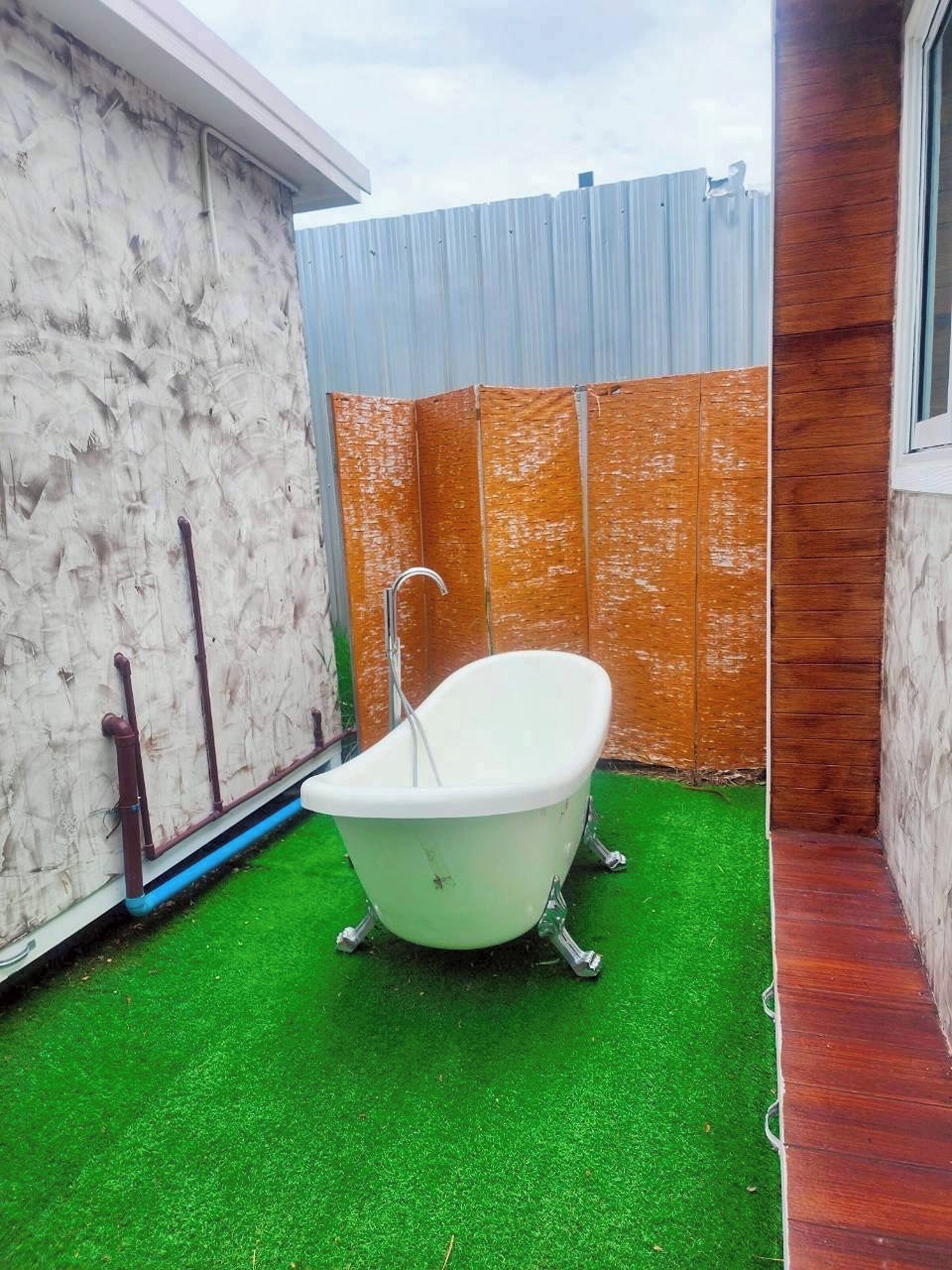 Una vasca da bagno bianca, indipendente, in un giardino con erba sintetica verde, una recinzione in metallo ondulato e un muro in cemento.