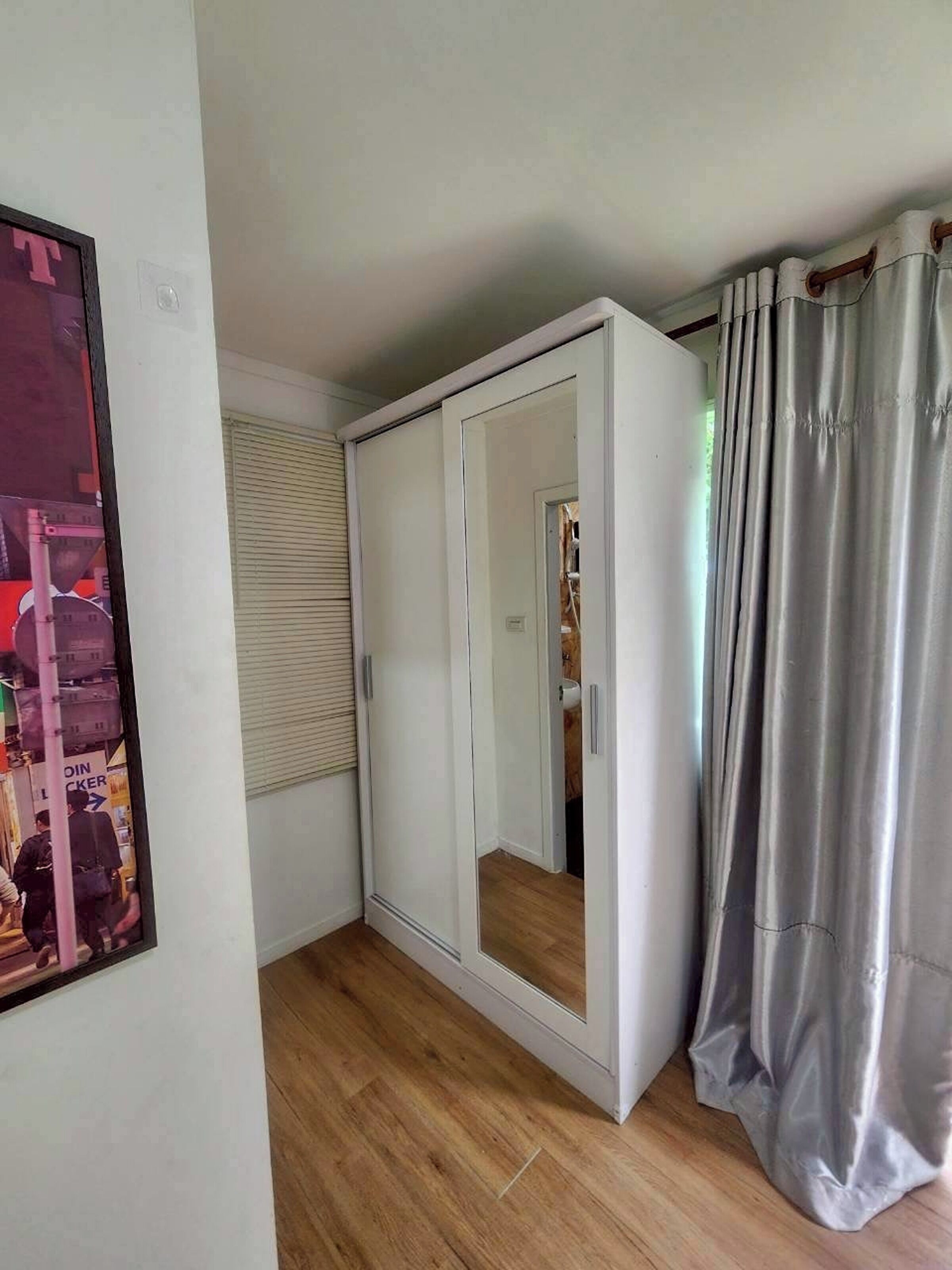 Une pièce avec une armoire blanche, un miroir, une fenêtre avec des stores et un rideau.
