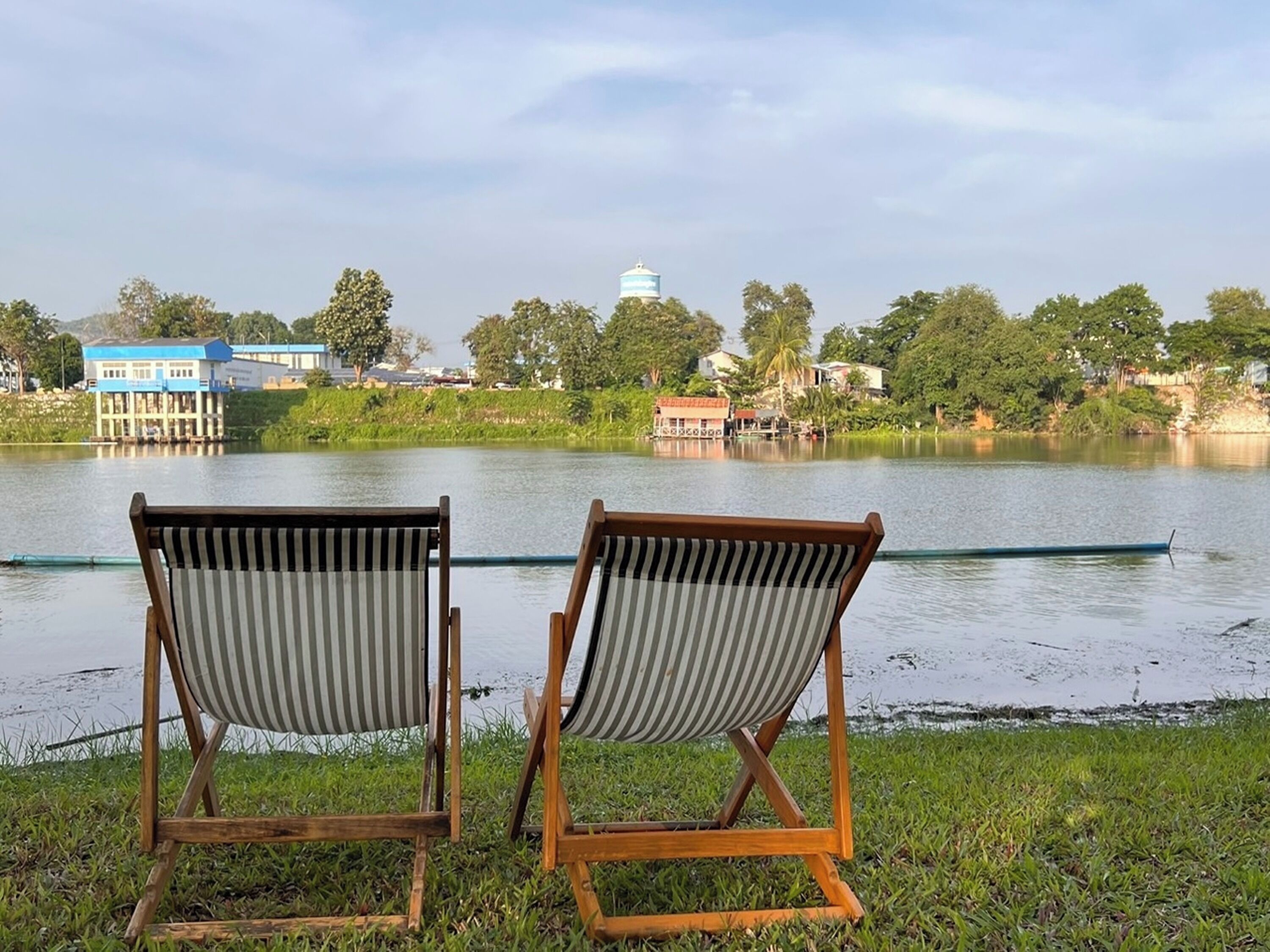 Two wooden chairs with striped cushions facing a calm lake, with buildings and trees in the background.