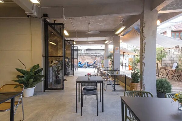 An outdoor cafe area with tables and chairs, potted plants, and a view of a street with parked scooters. - Nap & Nomad Hostel Tha Phae Gate (Chiang Mai)