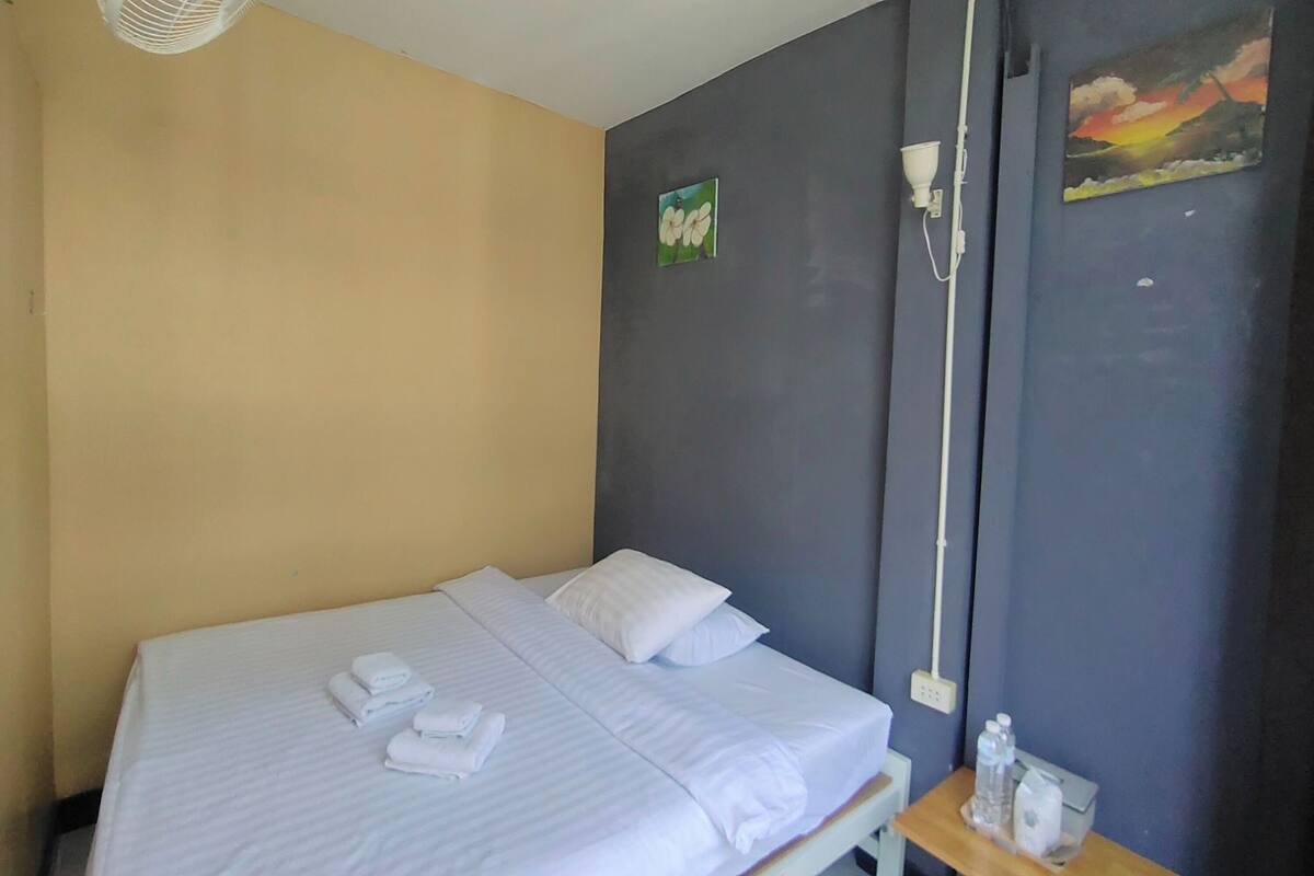 A neatly made bed with white linens and pillows, a bedside table with a water bottle and a tissue box, a wall-mounted light fixture, and a small painting of flowers.