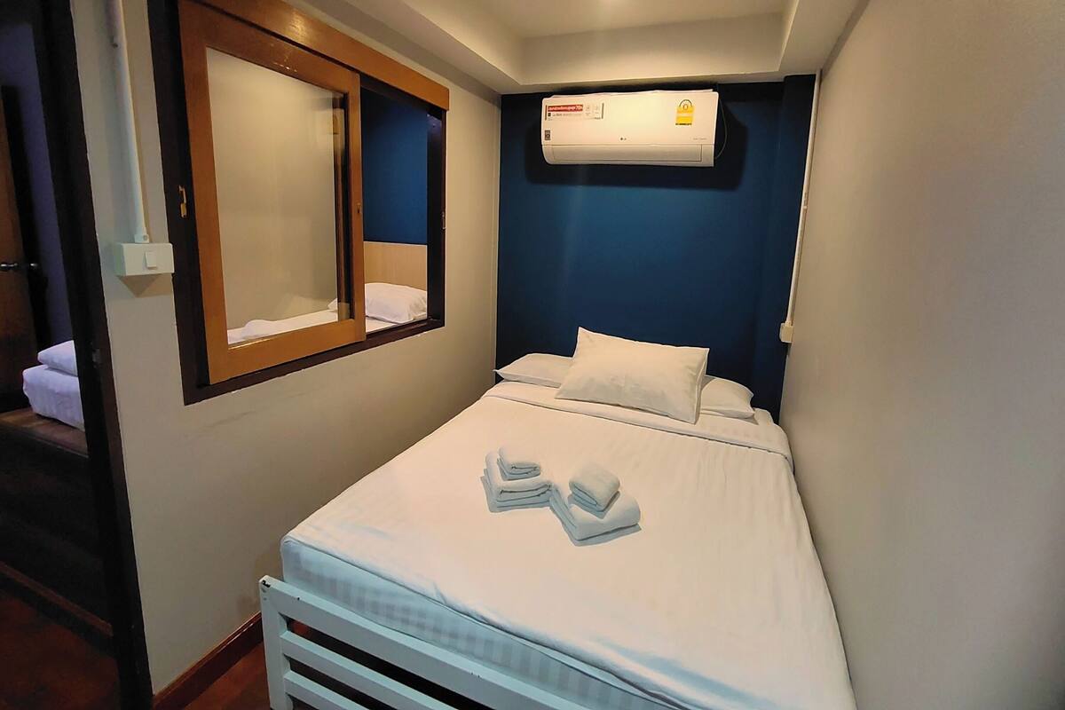 A small, neatly made bed with white linens and towels, a window with a wooden frame, and an air conditioning unit mounted on the wall.