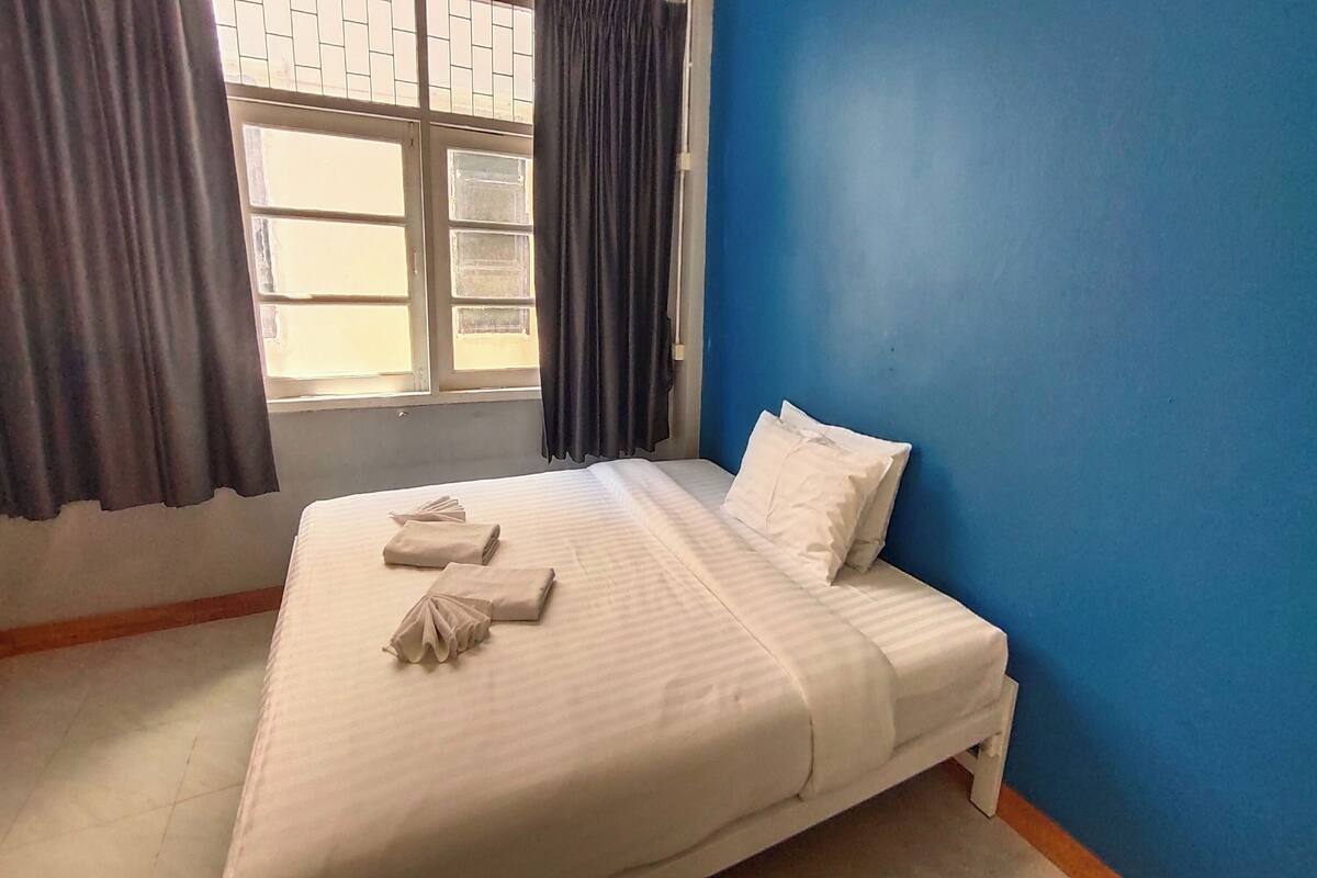 A neatly made bed with white linens and towels, a window with curtains, and a blue wall.