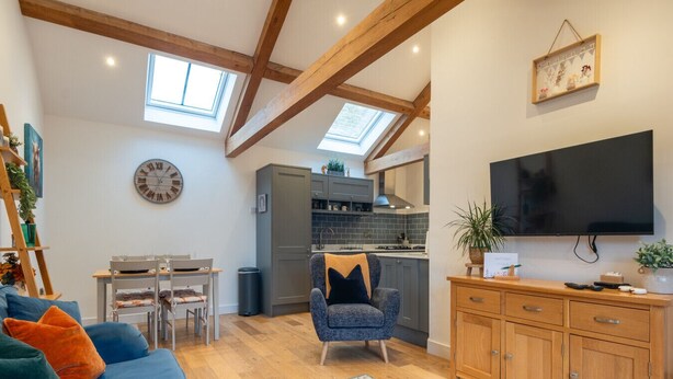 A modern living room with a blue sofa, a wooden dining table, and a kitchen in the background. - Beautiful 2 Bedroom Barn in Cerney Wick (Cerney Wick)