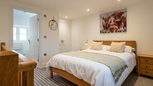 A bedroom with a wooden bed, a nightstand, a wall-mounted lamp, and a door leading to another room. - The Alehouse Retreat, The Crown Inn (Cerney Wick)