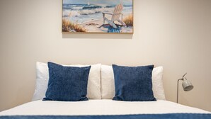 A neatly made bed with white and blue bedding, a painting of a beach scene above it, and a bedside lamp on the right. - Beautiful One Bedroom Cottage in Cerney Wick (Cerney Wick)