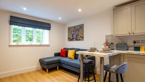 A modern kitchen with a dining area, featuring a dark grey sectional sofa, a marble-top table, and a small bar stool. - Beautiful One Bedroom Cottage in Cerney Wick (Cerney Wick)