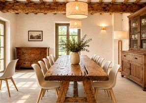 Un comedor rústico con una mesa de madera, sillas blancas, un jarrón con follaje y un armario de madera.