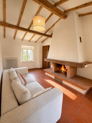 A living room with a fireplace, wooden beams, a sofa, and a window. - Tenuta Seriole (Poppi)