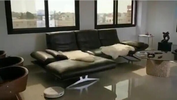 A modern living room with a black leather sofa, a white throw blanket, and a coffee table. There are large windows with a view of buildings outside. - thiego'sapparthotel (Lomé)