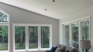 A modern living room with a grey sofa, a black armchair, and a floor lamp. Large windows offer a view of a forested area. - Large home with hot tub in serene wooded setting (Erie)