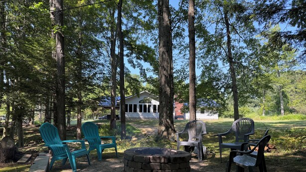 A forested campsite with a stone fire pit, several Adirondack chairs, and a covered picnic table. - Large home with hot tub in serene wooded setting (Erie)