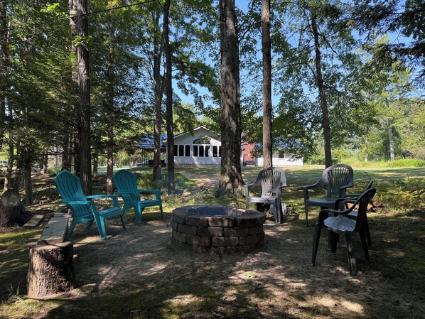 A forested campsite with a stone fire pit, several Adirondack chairs, and a covered picnic table. - Large home with hot tub in serene wooded setting (Erie)