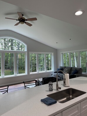 A modern kitchen with a large island, a ceiling fan, and large windows offering a view of a forest. - Large home with hot tub in serene wooded setting (Erie)