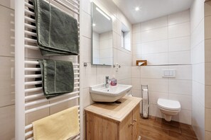 Ein Badezimmer mit einem weißen Waschbecken, einem Spiegel und einer Toilette. Handtücher befinden sich an einem Wäscheständer und es gibt einen Holzschrank.