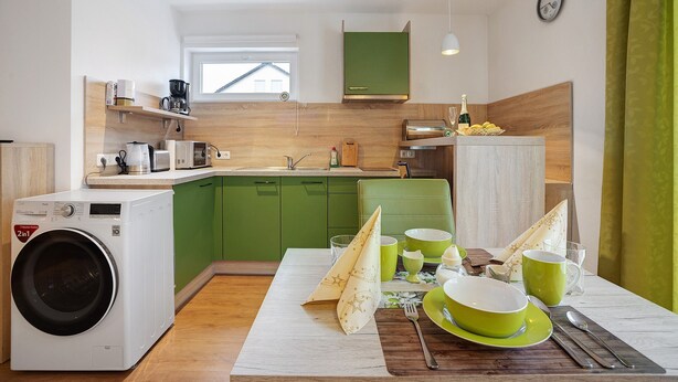A compact kitchen with a washing machine, a dining table set with green dishes, and a microwave. - Holiday Apartment Am Wiesenweg with Private Terrace and Wi-Fi (Mauerstetten)