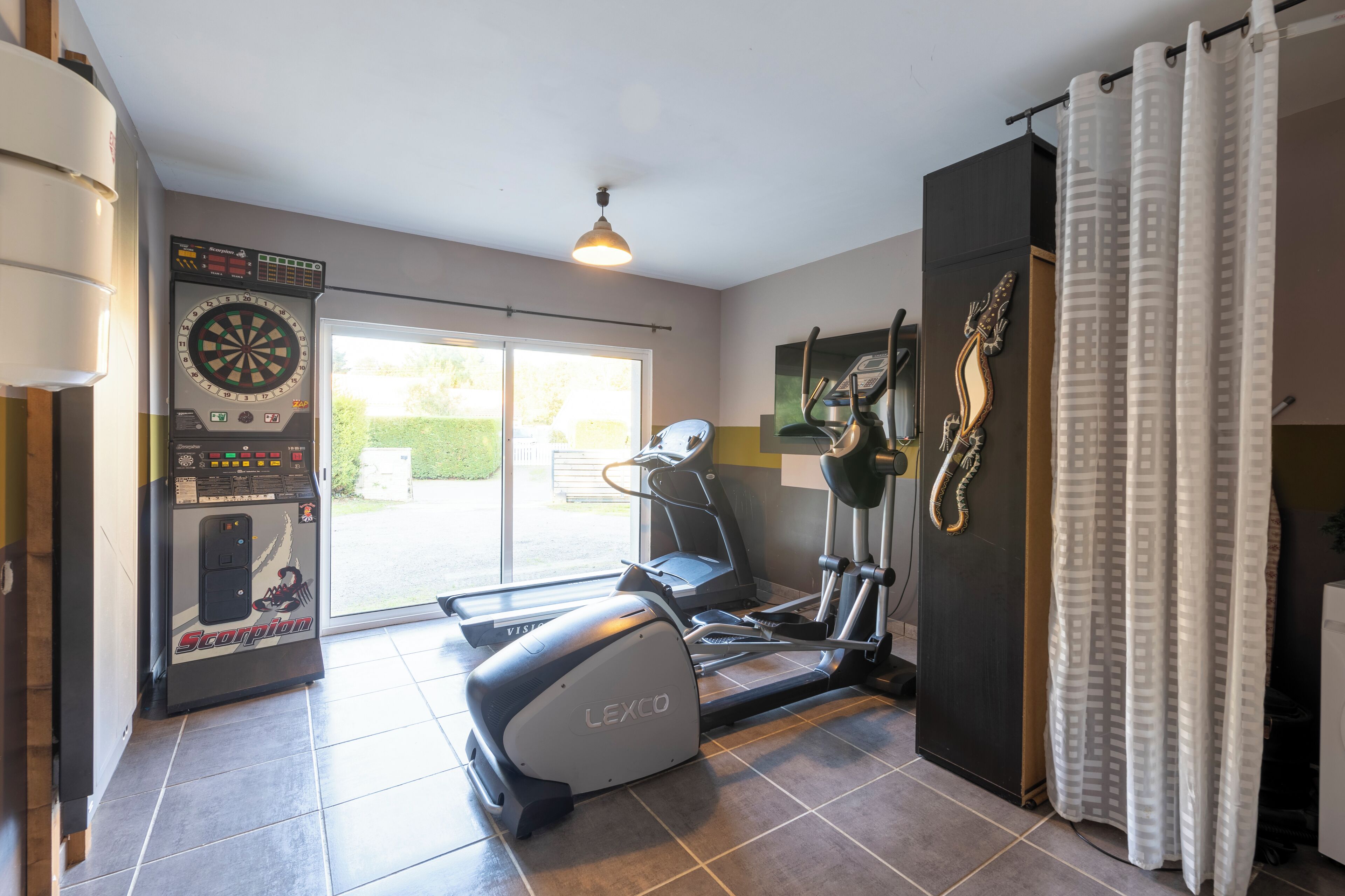 A home gym with a Lexco elliptical, a treadmill, a stationary bike, and a dartboard.
