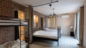 Een slaapzaal met stapelbedden, een bakstenen muur, een houten vloer, een plafondventilator en een raam met gordijnen.