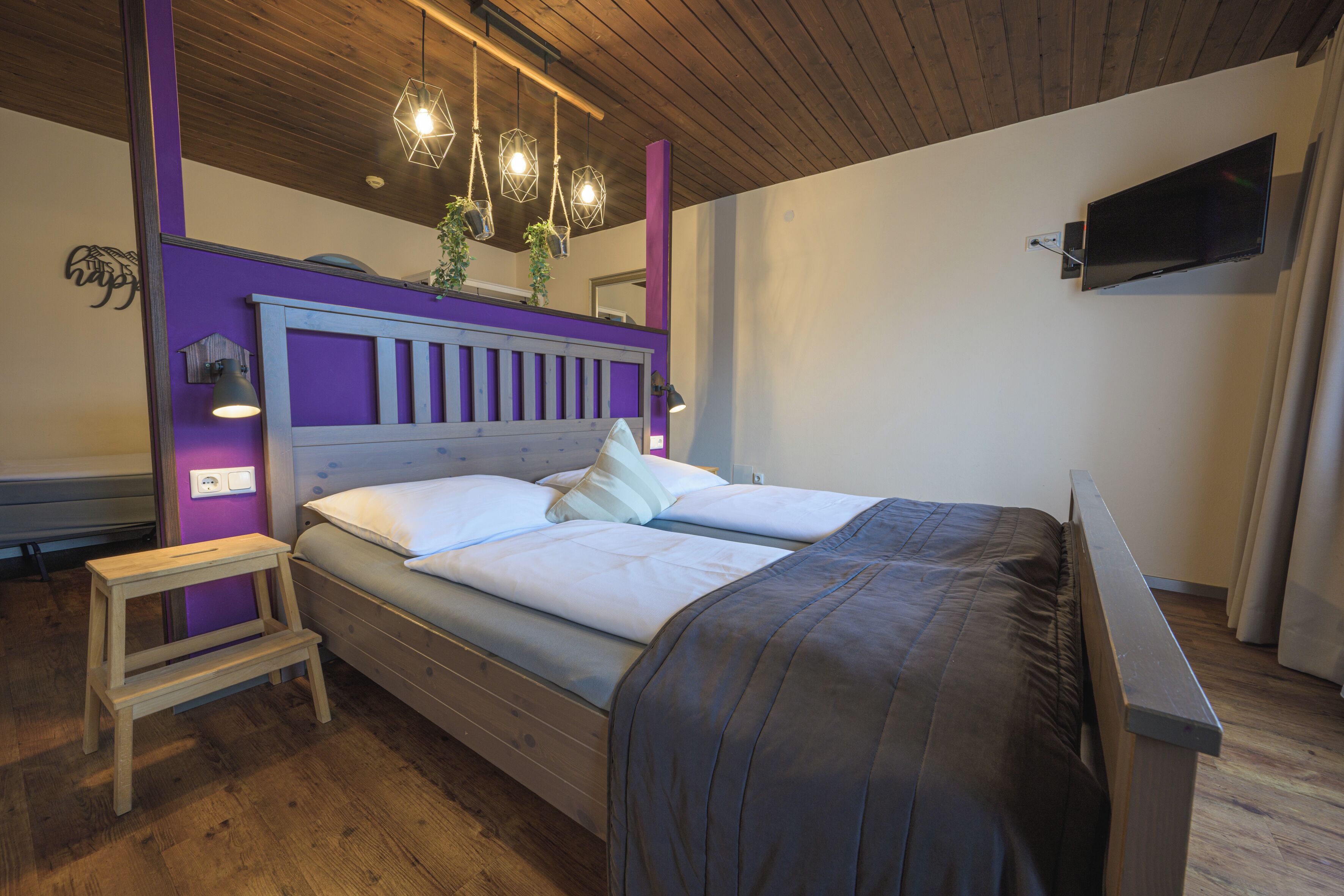A bedroom with a wooden floor, a bed with a headboard, a nightstand, a TV mounted on the wall, and a hanging light fixture.