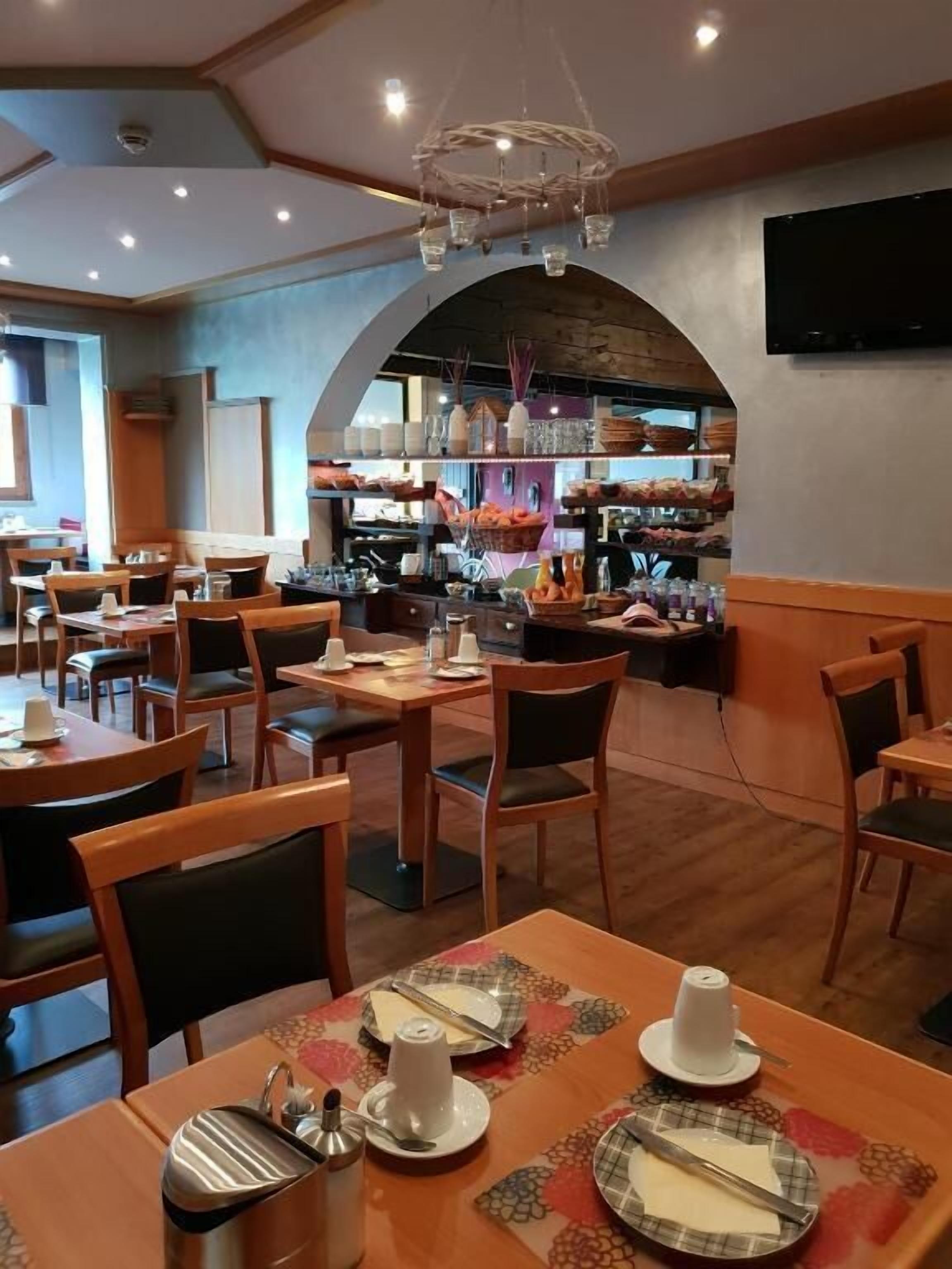 A restaurant with wooden tables and chairs, a bar area with shelves stocked with bottles, and a ceiling with recessed lighting.