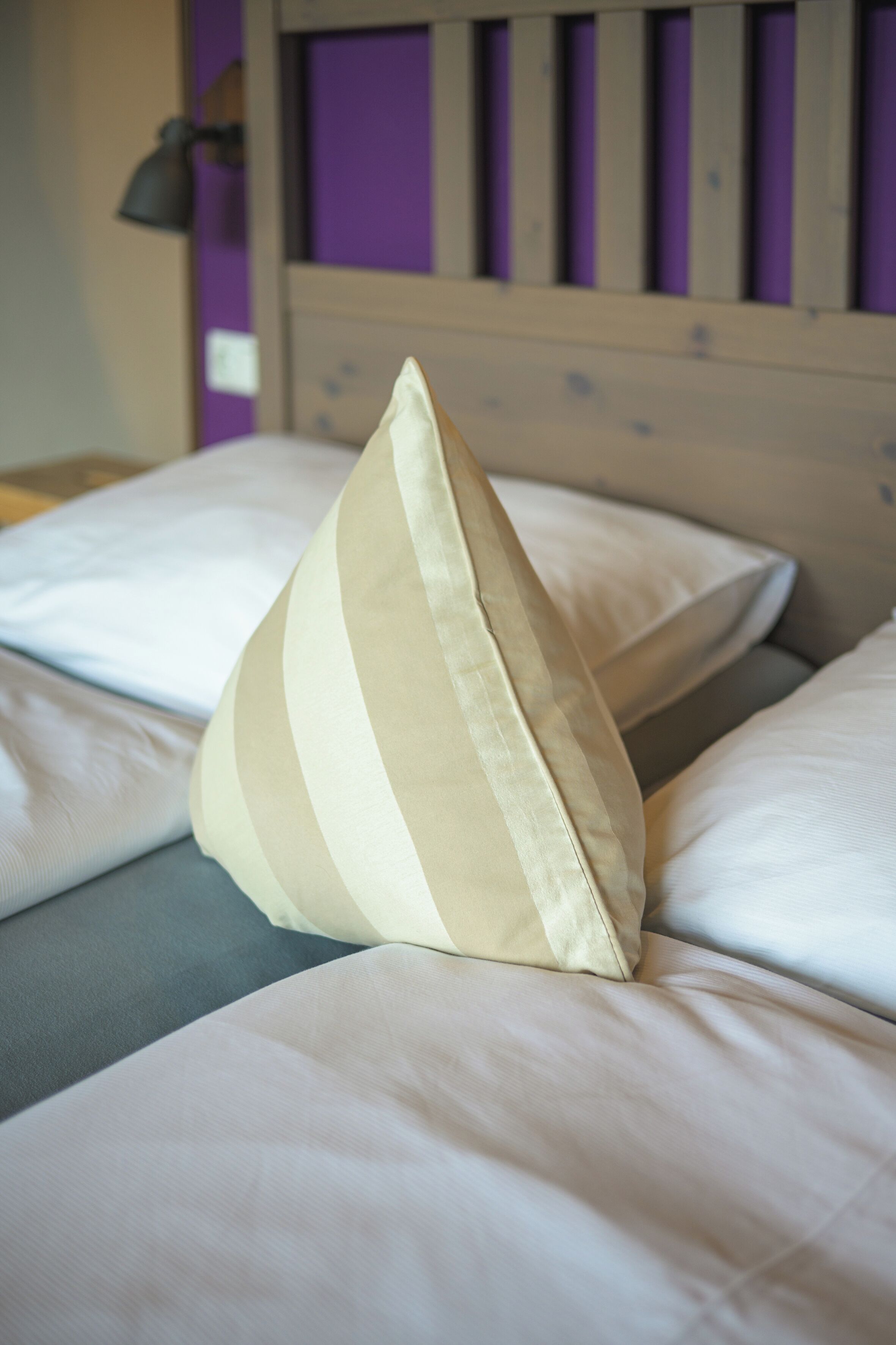 Ein Bett mit weißen und beigen Kissen, einem hölzernen Kopfteil und einer violetten Wand im Hintergrund.
