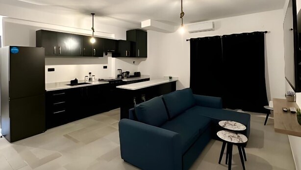 Una sala moderna con un sofá azul, una mesita redonda y una cocina con armarios negros y un refrigerador.
