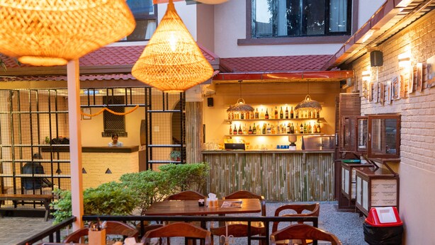 An outdoor dining area with wooden tables and chairs, a bar with shelves of bottles, and a brick wall with framed pictures. - Yangchhen Peace Home Bed & Breakfast (Kathmandu)