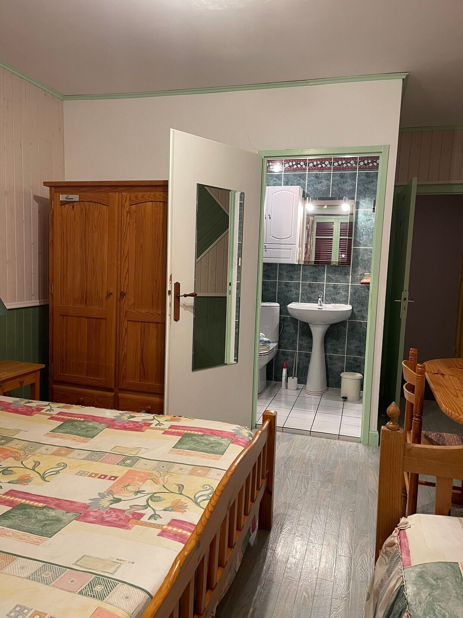 Une chambre à coucher avec une armoire en bois, un lit recouvert d’une courtepointe à motifs floraux, une chaise en bois et une salle de bain avec un lavabo et un miroir.