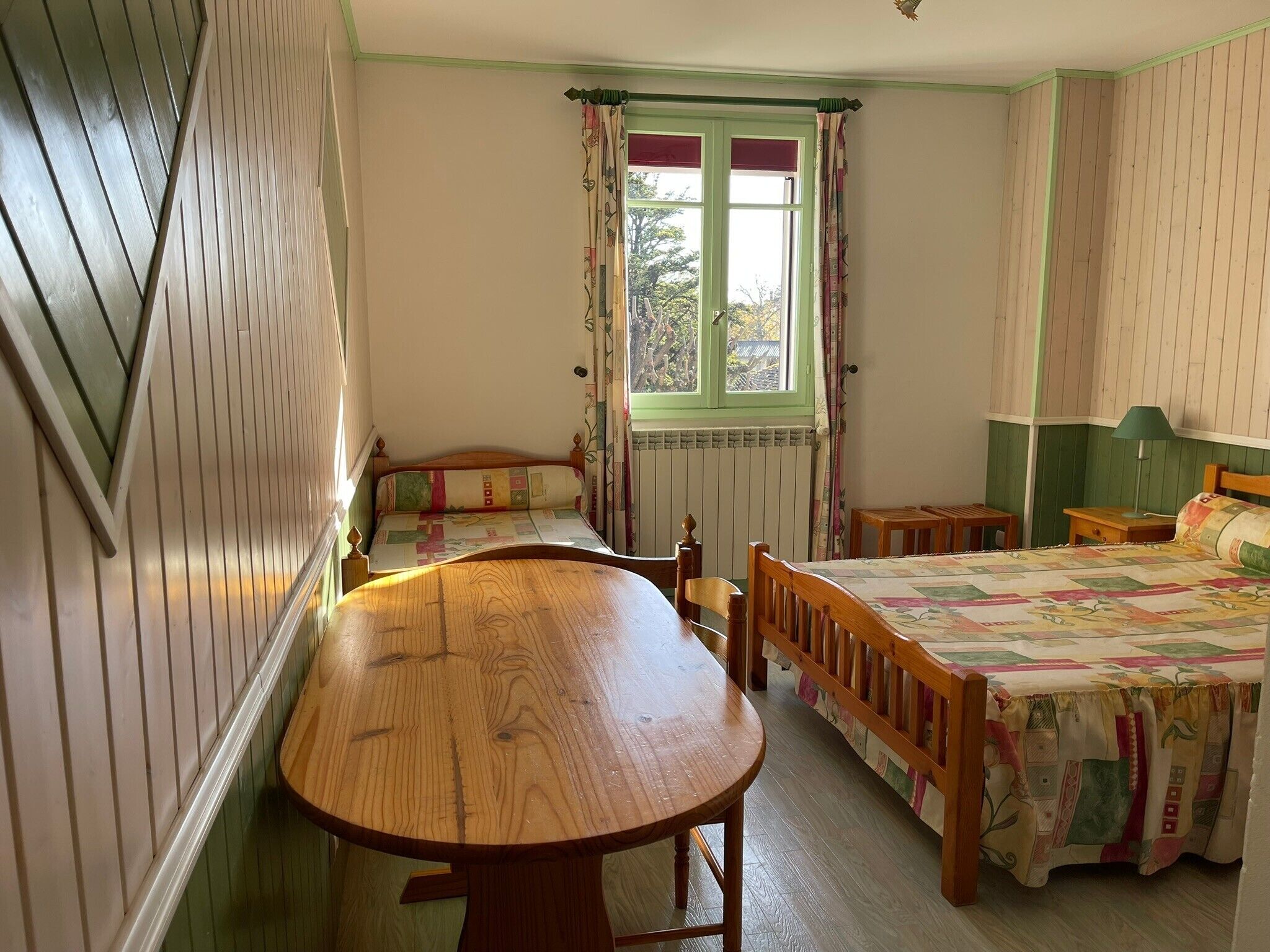 A room with a wooden table, a bed with a quilt, a window with curtains, and a radiator.