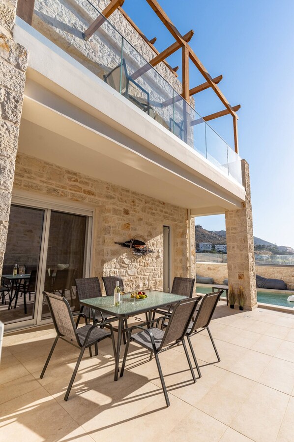 A modern outdoor dining area with a glass table and chairs, a stone wall, and a view of the sea. - Armonia Suite (Panormo)