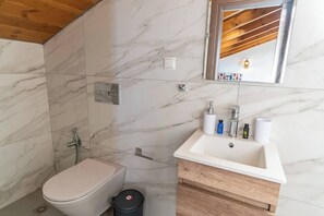 A modern bathroom with a marble floor, a white toilet, a wooden vanity with a sink, and a shower. - Armonia Suite (Panormo)