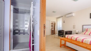 A hotel room with a bed, a wooden bench, a chair, a desk, a mirror, and a shower area. - Guest Room - La Rochelle (Virson)