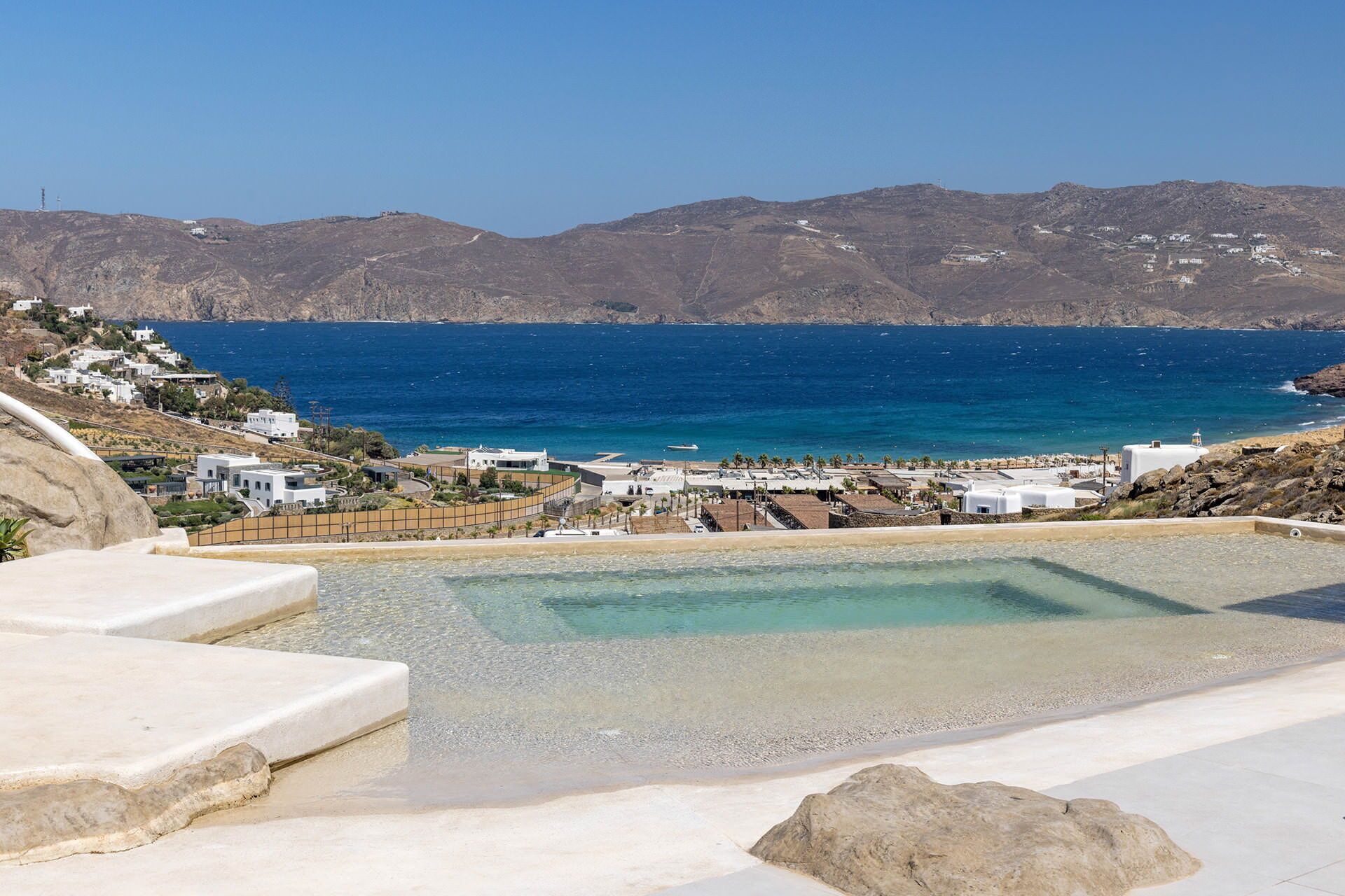 A coastal resort with a clear view of the sea, a swimming pool, and white buildings.
