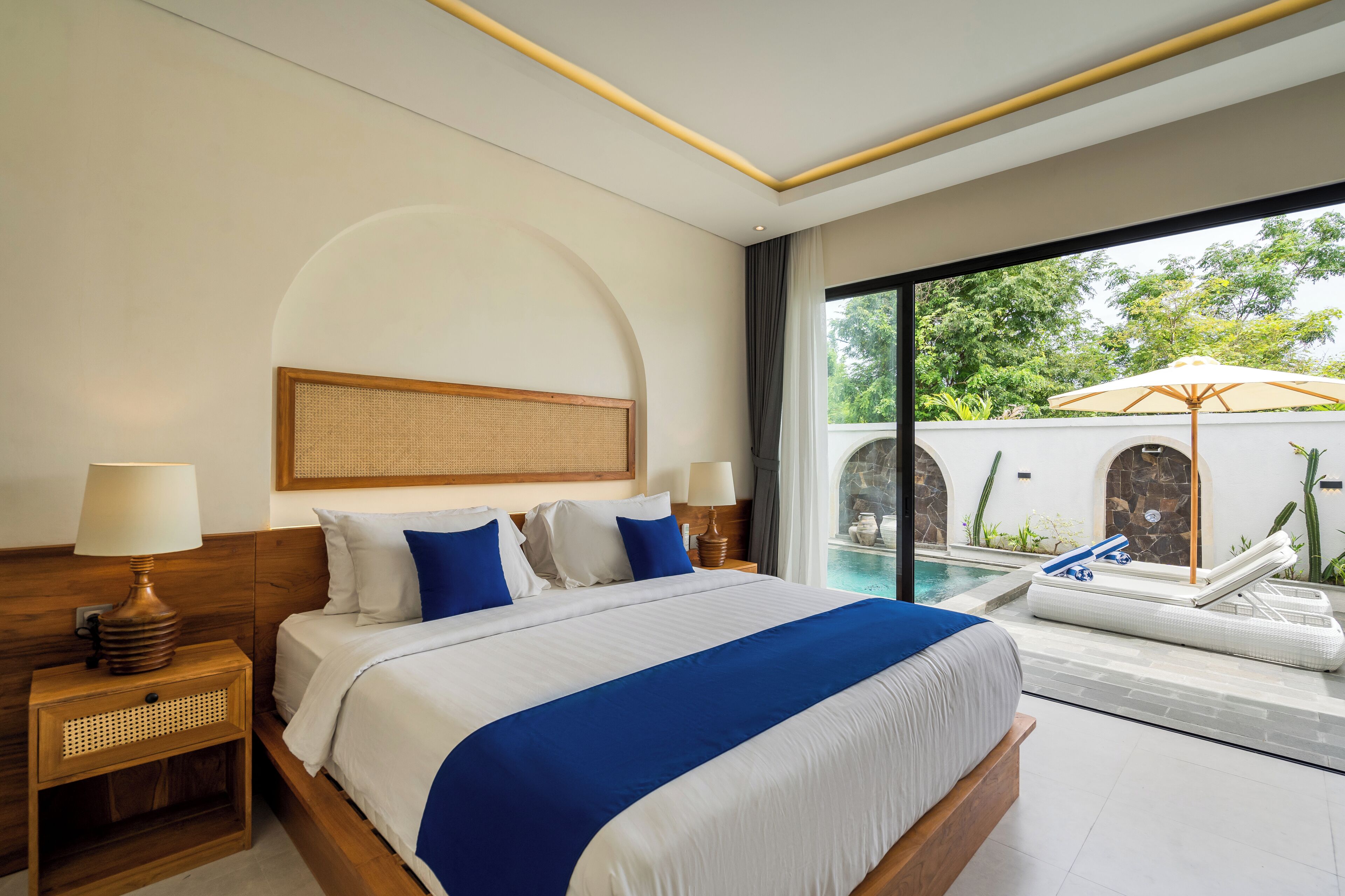 A hotel room with a large bed, two bedside tables, a window with a view of a pool and greenery, and a ceiling with a linear light fixture.