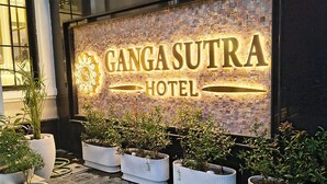 Exterior del hotel Ganga Sutra, con un llamativo letrero iluminado, plantas en macetas y pavimento adoquinado.