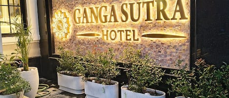 The exterior of the Ganga Sutra Hotel with a prominent illuminated sign, potted plants, and a tiled pavement.