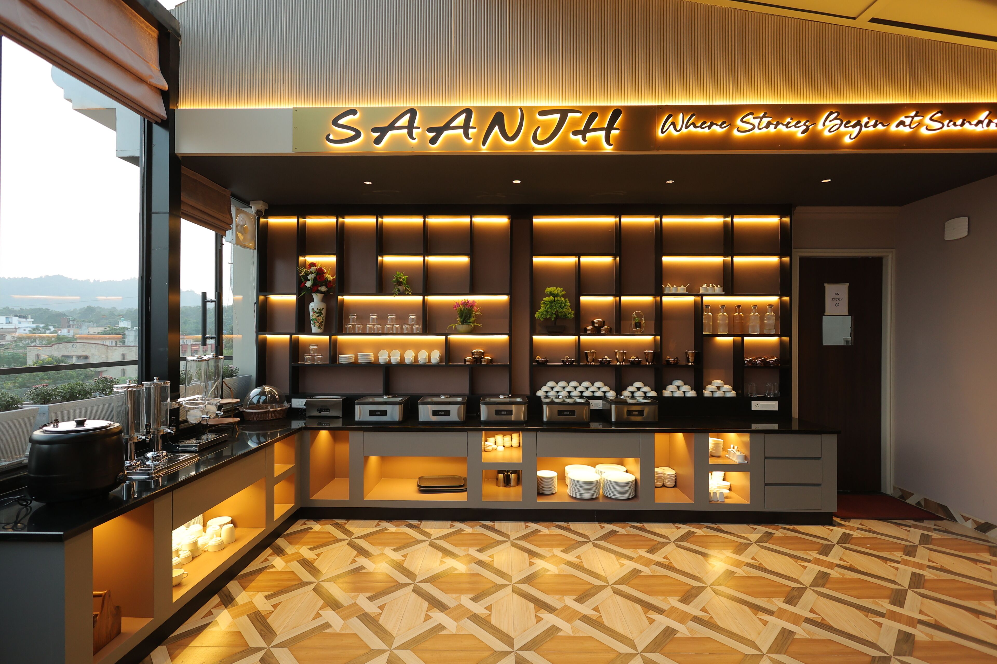 A modern hotel bar with a sign reading "SAANJH Where Stories Begin at Sandhi". It features a geometric patterned floor, a variety of glassware, and a view of the city through large windows.