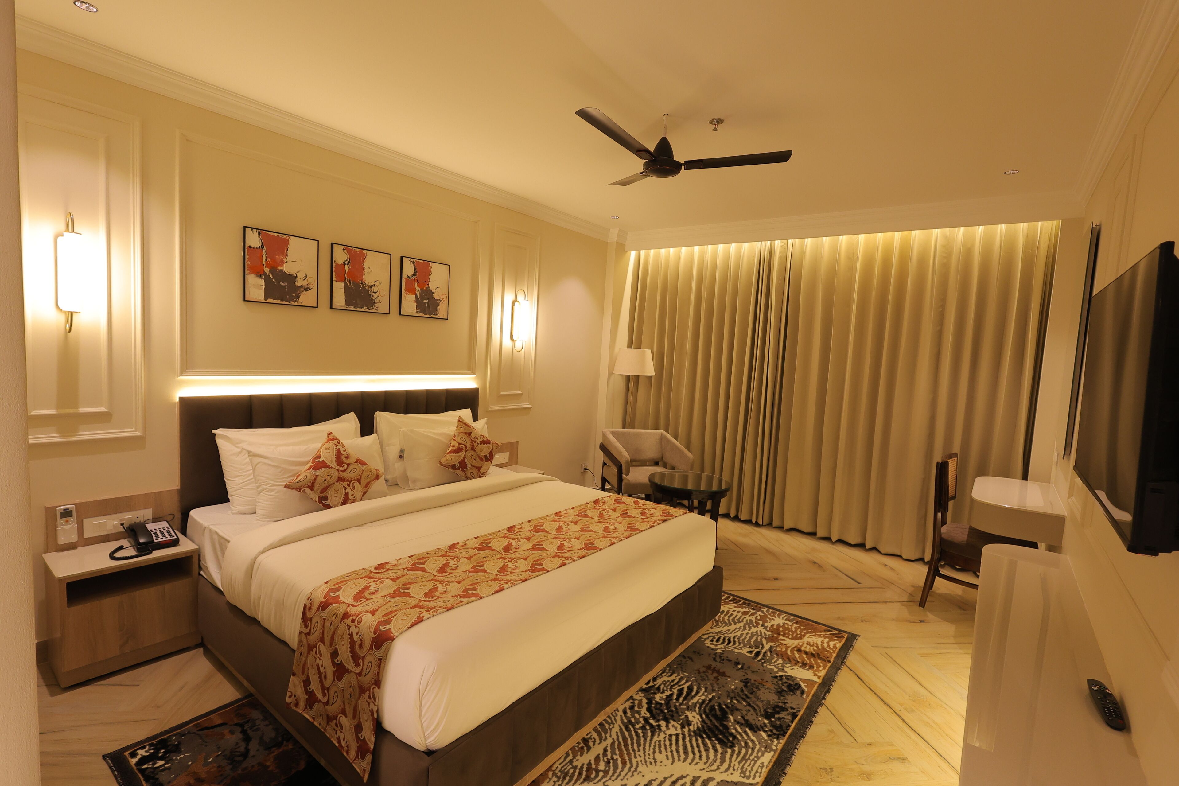 A hotel room with a large bed, bedside tables, a desk, a chair, a television, and a ceiling fan.